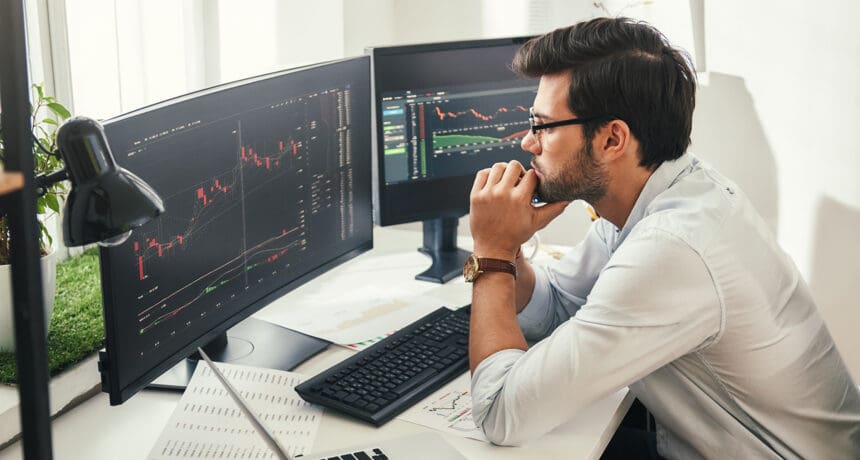 Successful trader. Back view of bearded stock market broker in eyeglasses analyzing data and graphs on multiple computer screens while sitting in modern office.